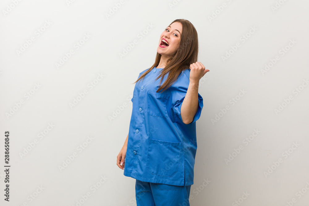 Young nurse woman against a white wall points with thumb finger away, laughing and carefree. Young nurse woman against a white wall points with thumb finger away, laughing and carefree.