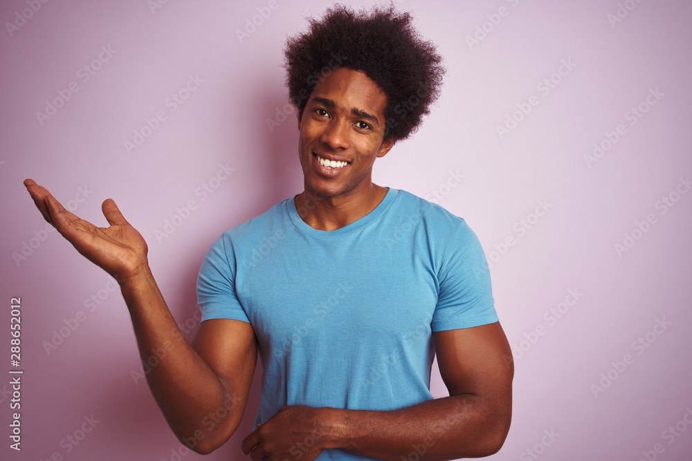 African american man with afro hair wearing blue t-shirt standing over isolated pink background smiling cheerful presenting and pointing with palm of hand looking at the camera. African american man with afro hair wearing blue t-shirt standing over isolated pink background smiling cheerful presenting and pointing with palm of hand looking at the camera.