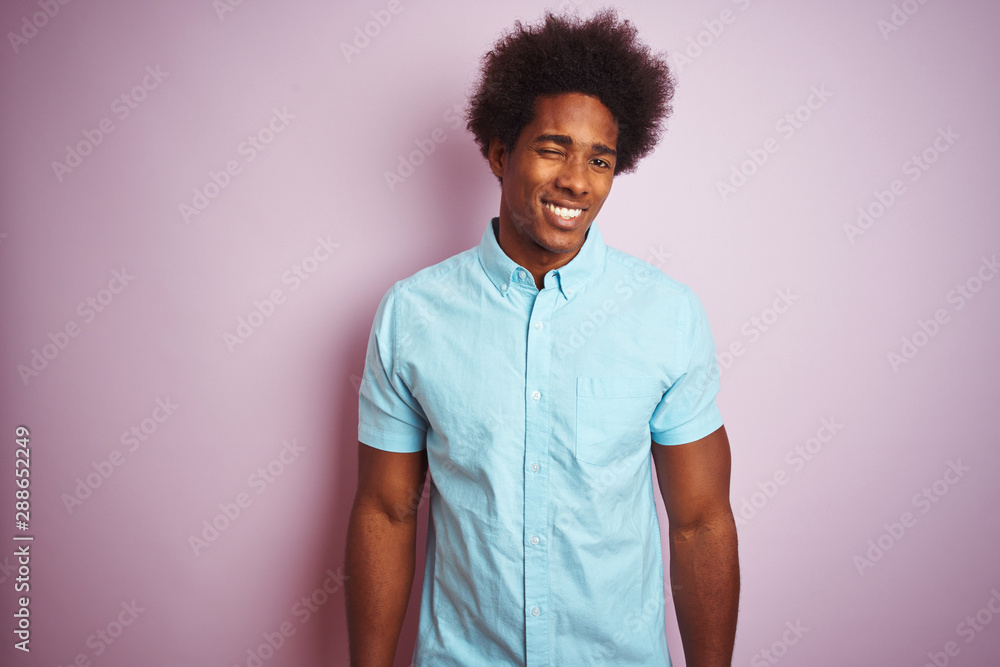 Young american man with afro hair wearing blue shirt standing over isolated pink background winking looking at the camera with sexy expression, cheerful and happy face. Young american man with afro hair wearing blue shirt standing over isolated pink background winking looking at the camera with sexy expression, cheerful and happy face.