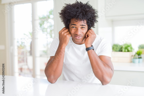 Schilderij op canvas Young african american man wearing casual white t-shirt sitting at home covering ears with fingers with annoyed expression for the noise of loud music
