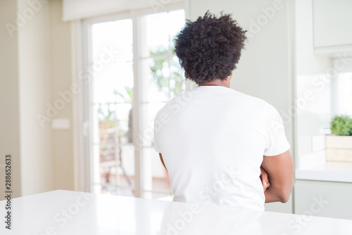 Young african american man wearing casual white t-shirt sitting at home standing backwards looking away with crossed arms