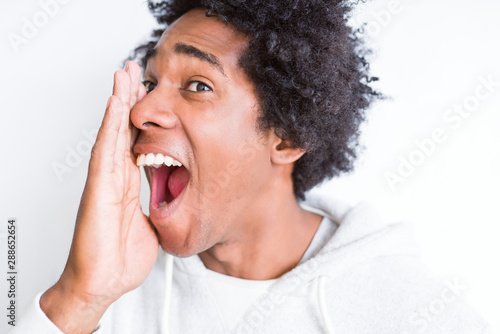 Handsome african american man with afro hair over white background shouting and screaming loud to side with hand on mouth. Communication concept.