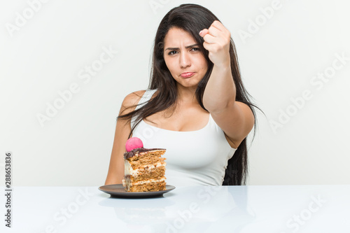 Young hispanic woman eating a cake showing fist to camera, aggressive facial expression.