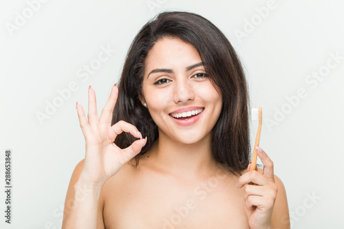 Young hispanic woman holding a toothbrush cheerful and confident showing ok gesture.