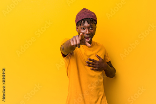 Young black man wearing rastas over yellow background points with thumb finger away, laughing and carefree.