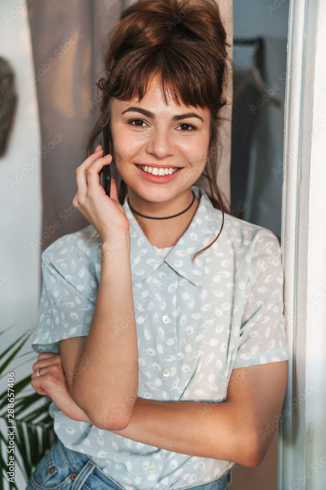 Cheerful pleased happy young beautiful woman talking by mobile phone indoors at home. Cheerful pleased happy young beautiful woman talking by mobile phone indoors at home.