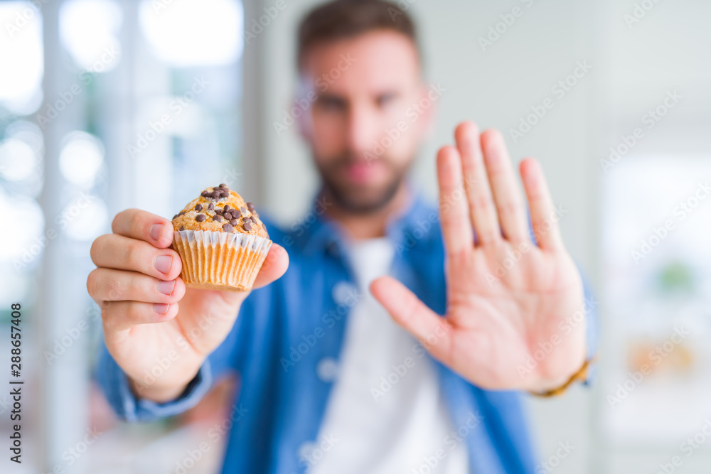 Handsome man eating chocolate chips muffin with open hand doing stop sign with serious and confident expression, defense gesture