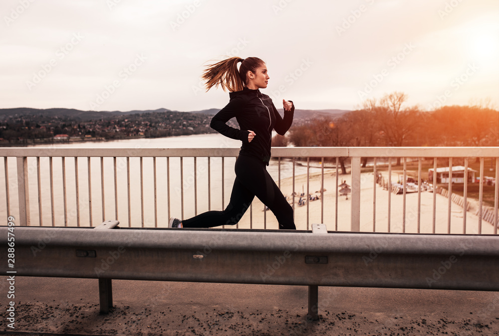 Young woman in black sports outfit running on the bridge in the city during sunset. Young woman in black sports outfit running on the bridge in the city during sunset.