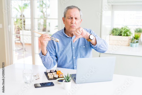 Handsome business senior man eating delivery sushi while working using laptop with angry face, negative sign showing dislike with thumbs down, rejection concept