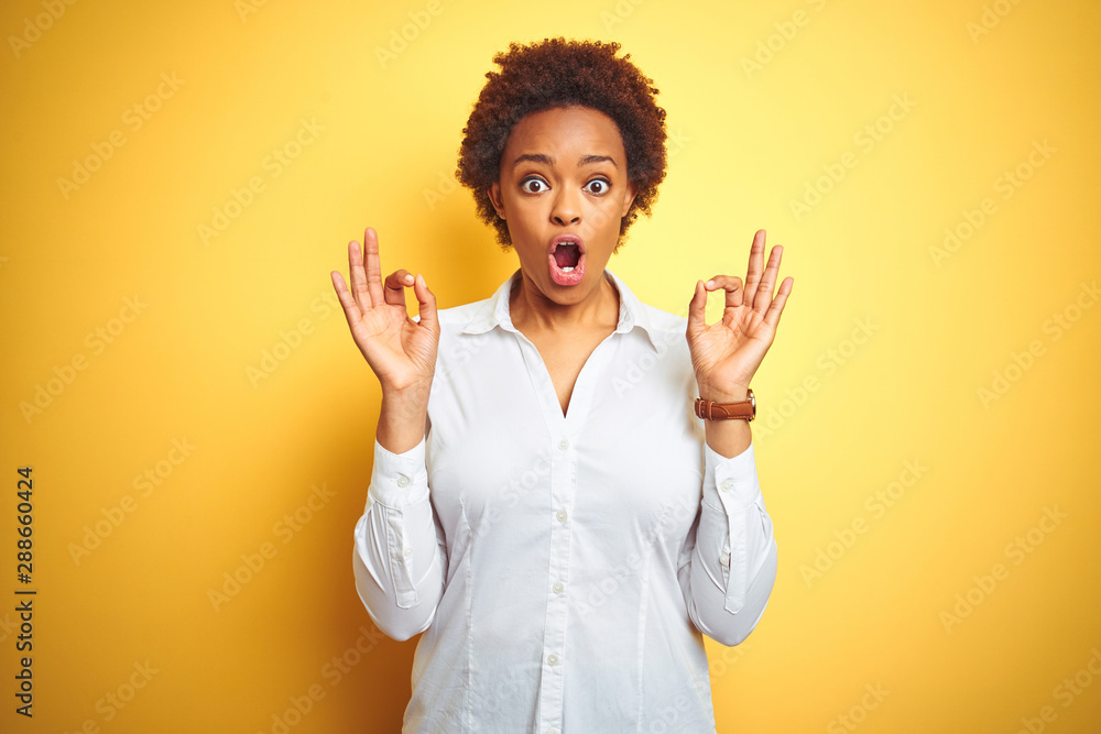 African american business woman over isolated yellow background looking surprised and shocked doing ok approval symbol with fingers. Crazy expression African american business woman over isolated yellow background looking surprised and shocked doing ok approval symbol with fingers. Crazy expression