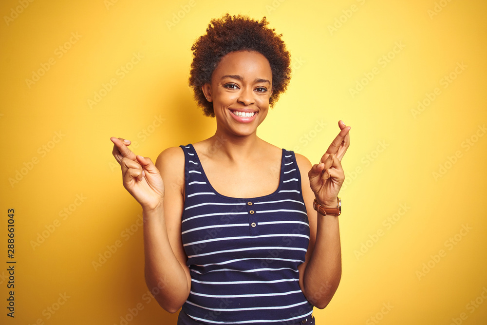Beauitul african american woman wearing summer t-shirt over isolated yellow background gesturing finger crossed smiling with hope and eyes closed. Luck and superstitious concept. Beauitul african american woman wearing summer t-shirt over isolated yellow background gesturing finger crossed smiling with hope and eyes closed. Luck and superstitious concept.