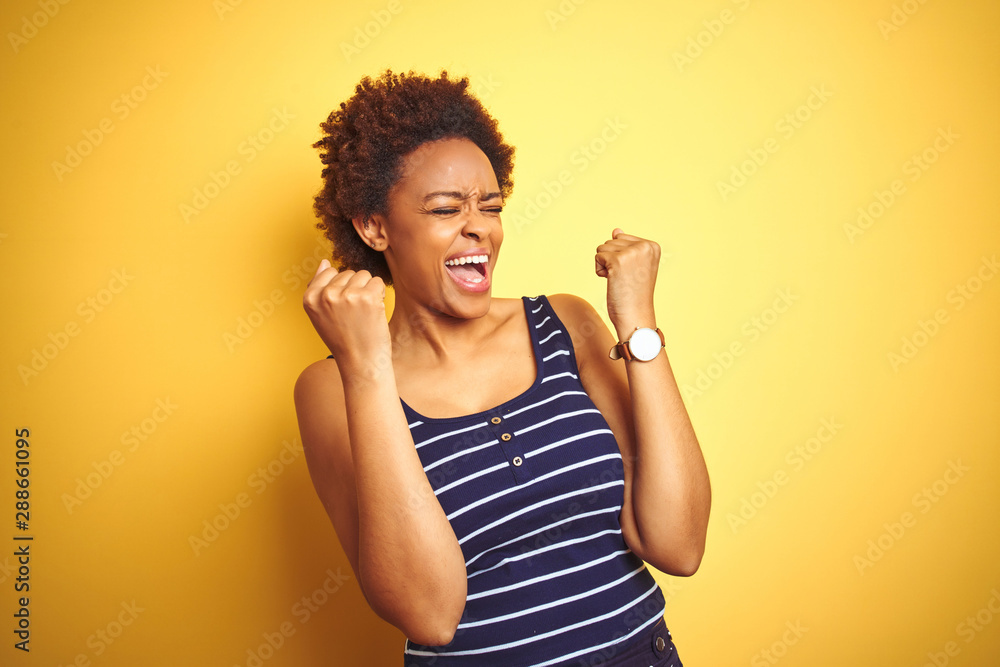 Beauitul african american woman wearing summer t-shirt over isolated yellow background celebrating surprised and amazed for success with arms raised and eyes closed. Winner concept. Beauitul african american woman wearing summer t-shirt over isolated yellow background celebrating surprised and amazed for success with arms raised and eyes closed. Winner concept.