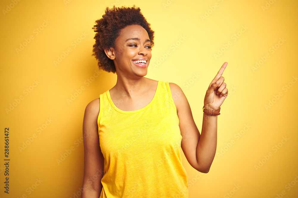 Beauitul african american woman wearing summer t-shirt over isolated yellow background with a big smile on face, pointing with hand and finger to the side looking at the camera. Beauitul african american woman wearing summer t-shirt over isolated yellow background with a big smile on face, pointing with hand and finger to the side looking at the camera.