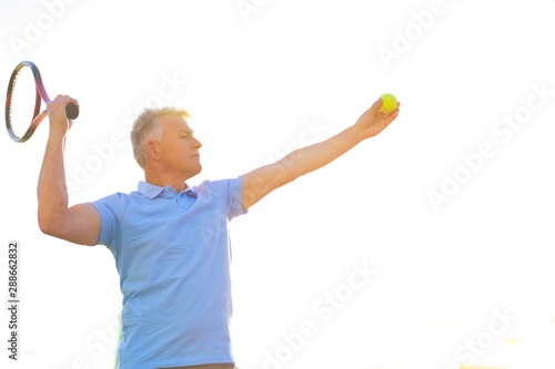 Confident mature man hitting tennis ball with racket on court against clear blue sky