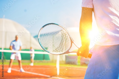 Active seniors playing tennis on sunny day at red court with yellow lens flare in background