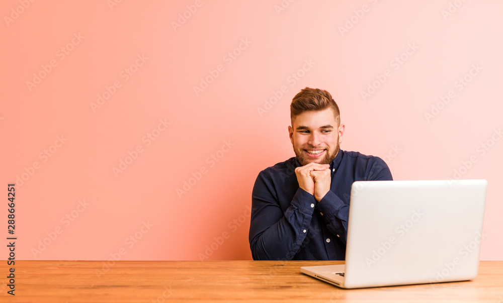 Young man working with his laptop keeps hands under chin, is looking happily aside. Young man working with his laptop keeps hands under chin, is looking happily aside.