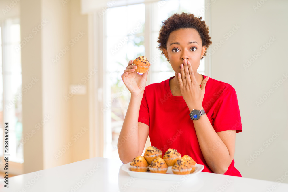 Young african american woman eating chocolate chips muffins cover mouth with hand shocked with shame for mistake, expression of fear, scared in silence, secret concept Young african american woman eating chocolate chips muffins cover mouth with hand shocked with shame for mistake, expression of fear, scared in silence, secret concept