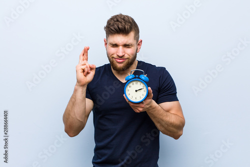 Young caucasian man holding alarm clock crossing fingers for having luck photo