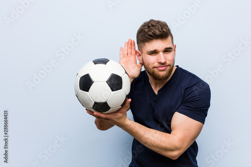 Young caucasian man holding a soccer ball trying to listening a gossip.