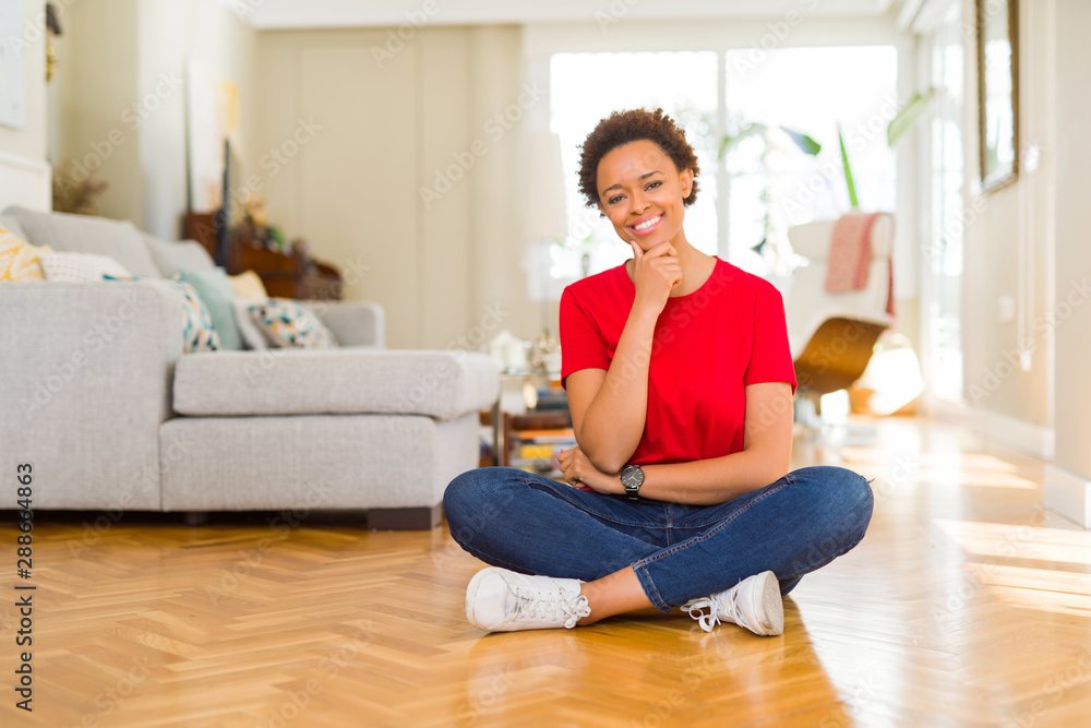 Young beautiful african american woman sitting on the floor at home looking confident at the camera with smile with crossed arms and hand raised on chin. Thinking positive. Young beautiful african american woman sitting on the floor at home looking confident at the camera with smile with crossed arms and hand raised on chin. Thinking positive.