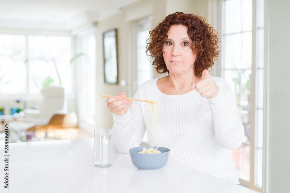 Senior woman eating asian noodles using chopsticks pointing with finger to the camera and to you, hand sign, positive and confident gesture from the front