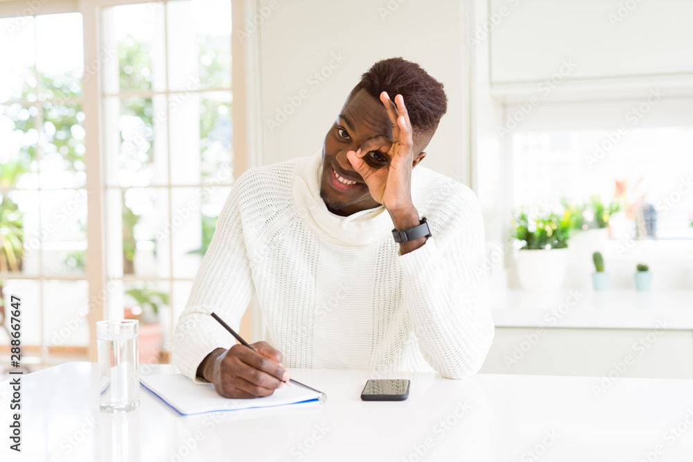African american student man writing on a paper using a pencil with happy face smiling doing ok sign with hand on eye looking through fingers African american student man writing on a paper using a pencil with happy face smiling doing ok sign with hand on eye looking through fingers