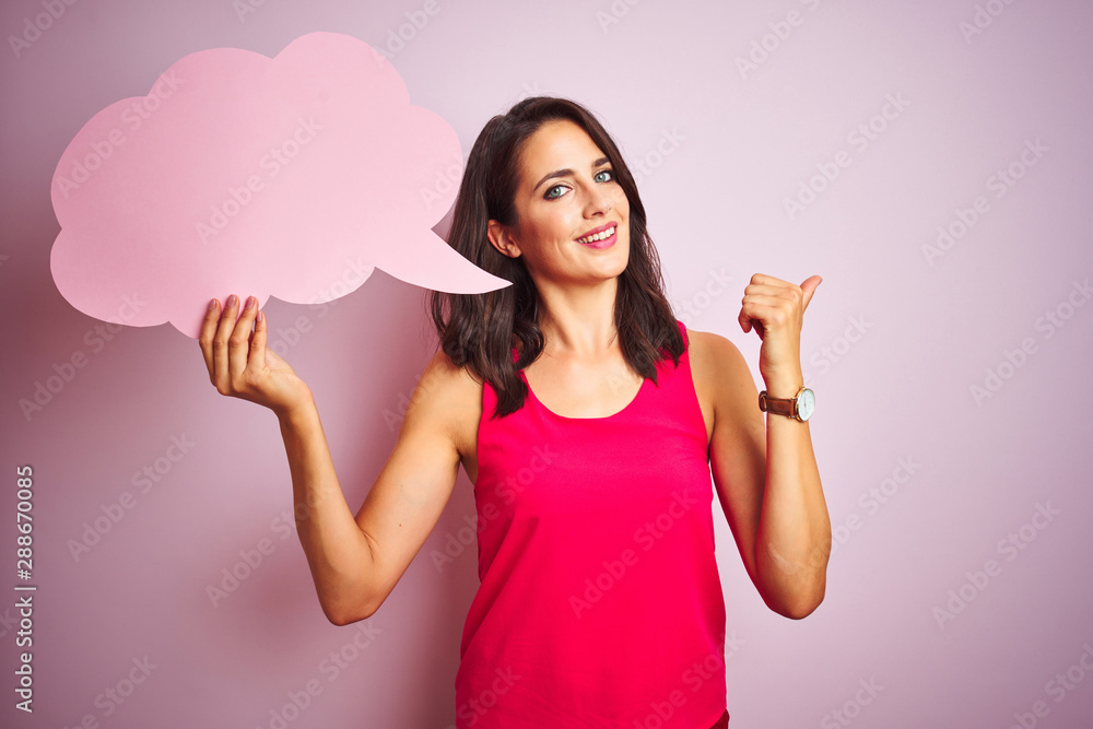 Beautiful woman holding speech bubble over pink isolated background pointing and showing with thumb up to the side with happy face smiling Beautiful woman holding speech bubble over pink isolated background pointing and showing with thumb up to the side with happy face smiling