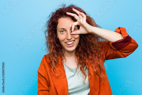 Young redhead elegant woman excited keeping ok gesture on eye.