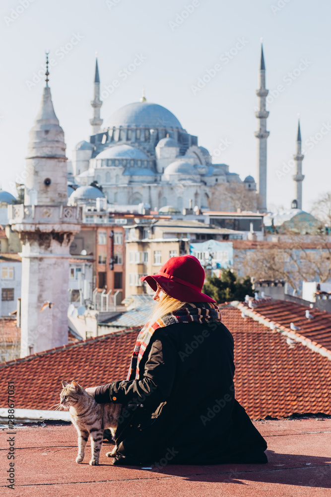 Obraz premium Blond woman stroking a cat on the roof with a view to the mosque, Istanbul, Turkey. Girl in a hat sits on the roof in Istanbul, sunny autumn day. Traveler girl walks through winter Istanbul.
