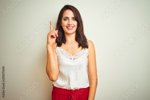 Young beautiful woman wearing t-shirt standing over white isolated background showing and pointing up with finger number one while smiling confident and happy.