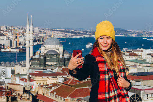 Blond woman makes photo on the phone on the roof of the Grand Bazaar, Istanbul, Turkey. Girl in a yellow hat takes a selfie on a sunny autumn day. Traveler girl walks through winter Istanbul.