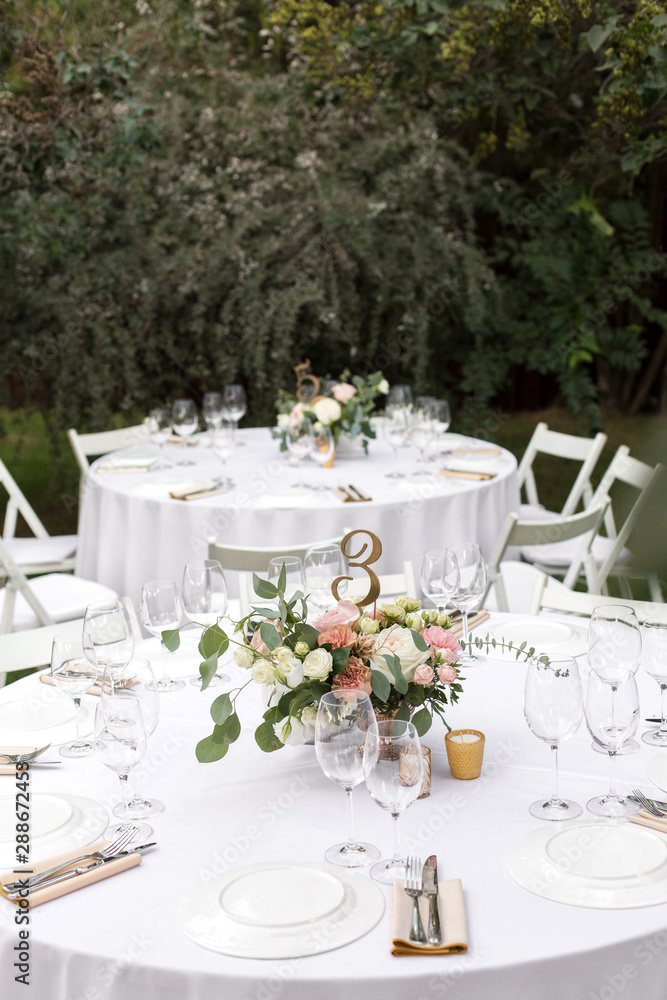 Fototapeta premium Wedding table setting decorated with fresh flowers in a brass vase. Wedding floristry. Banquet table for guests outdoors with a view of green nature. Bouquet with roses, eustoma and eucalyptus leaves