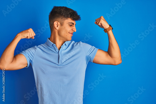 Young indian man wearing casual polo standing over isolated blue background showing arms muscles smiling proud. Fitness concept.