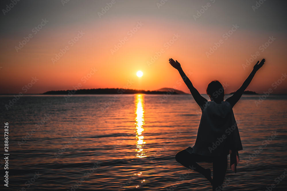Happy woman standing on one leg cheering on the beach, shot at sunset during summer vacation in Greece
