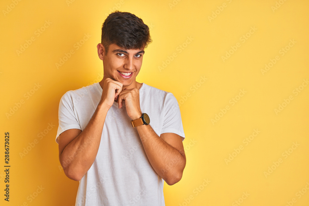 Young indian man wearing white t-shirt standing over isolated yellow background laughing nervous and excited with hands on chin looking to the side Young indian man wearing white t-shirt standing over isolated yellow background laughing nervous and excited with hands on chin looking to the side
