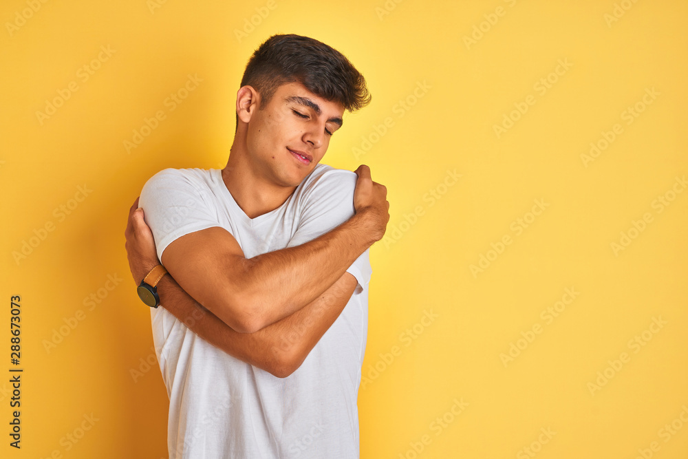 Young indian man wearing white t-shirt standing over isolated yellow background Hugging oneself happy and positive, smiling confident. Self love and self care Young indian man wearing white t-shirt standing over isolated yellow background Hugging oneself happy and positive, smiling confident. Self love and self care