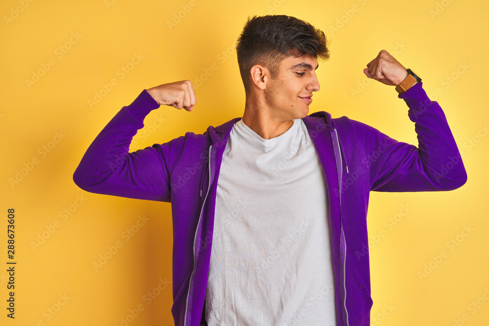 Young indian man wearing purple sweatshirt standing over isolated yellow background showing arms muscles smiling proud. Fitness concept. Young indian man wearing purple sweatshirt standing over isolated yellow background showing arms muscles smiling proud. Fitness concept.