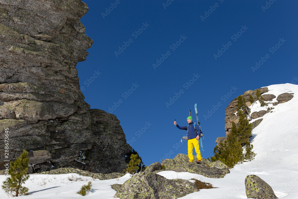 young guy takes a selfie on the phone in the mountains standing with skis behind his back. Young active man ski touring and staying at mountain peak background. ski tour