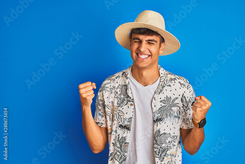 Indian man on vacation wearing hawiaian shirt summer hat over isolated blue background very happy and excited doing winner gesture with arms raised, smiling and screaming for success