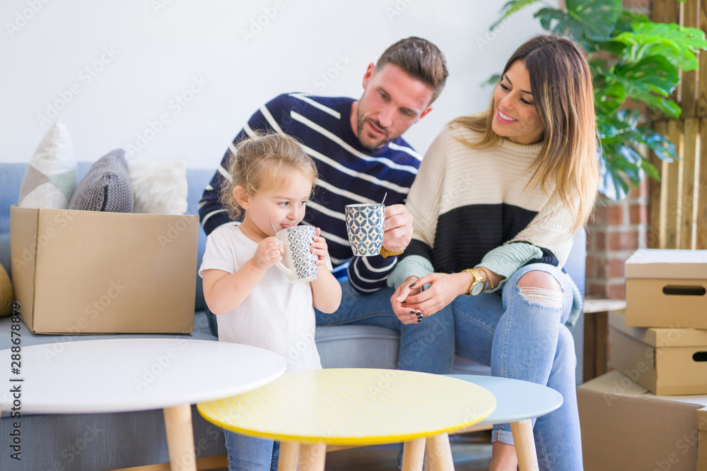 Beautiful family, parents sitting on the sofa drinking coffee looking his kid playing at new home around cardboard boxes Beautiful family, parents sitting on the sofa drinking coffee looking his kid playing at new home around cardboard boxes