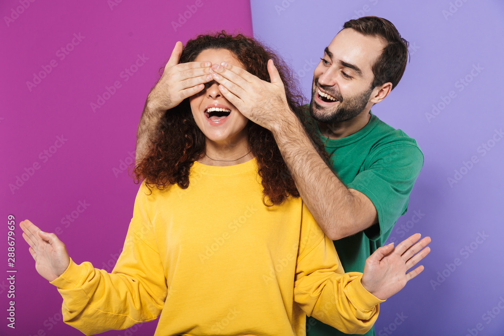 Photo of happy man in colorful clothing smiling and covering woman's eyes standing behind her Photo of happy man in colorful clothing smiling and covering woman's eyes standing behind her