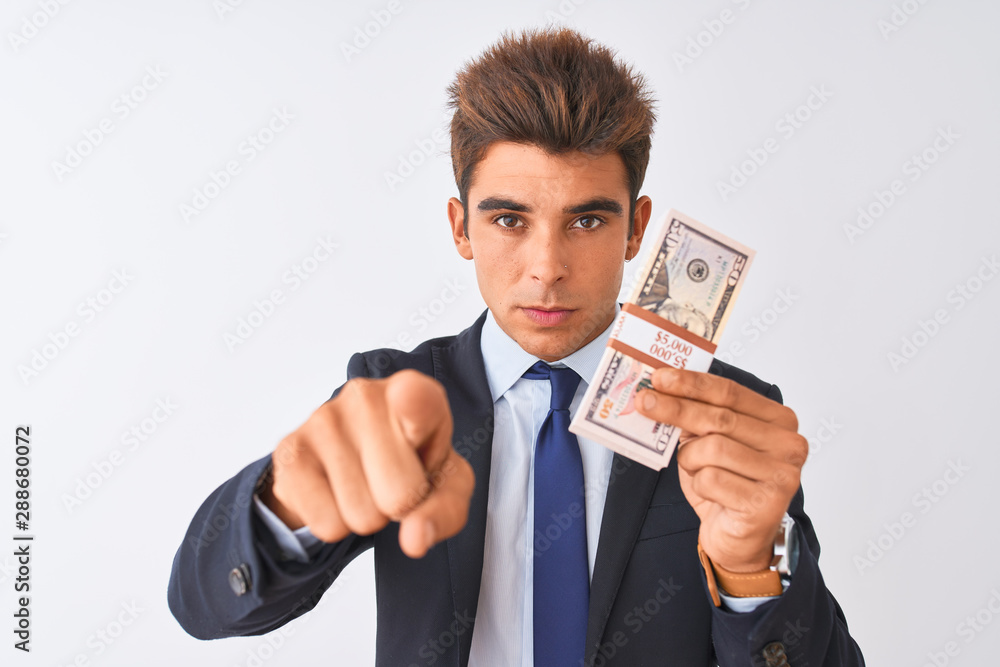 Young handsome businessman wearing suit holding dollars over isolated white background pointing with finger to the camera and to you, hand sign, positive and confident gesture from the front Young handsome businessman wearing suit holding dollars over isolated white background pointing with finger to the camera and to you, hand sign, positive and confident gesture from the front