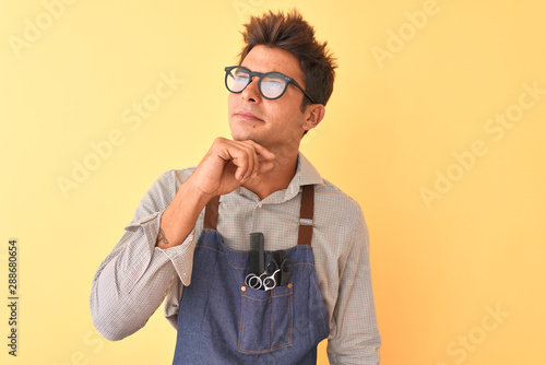 Young handsome hairdresser man wearing apron and glasses over isolated yellow background with hand on chin thinking about question, pensive expression. Smiling with thoughtful face. Doubt concept.