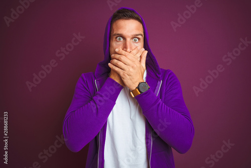 Young fitness man wearing casual sports sweatshirt and hood over purple isolated background shocked covering mouth with hands for mistake. Secret concept.