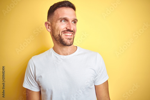 Young handsome man wearing casual white t-shirt over yellow isolated background looking away to side with smile on face, natural expression. Laughing confident.