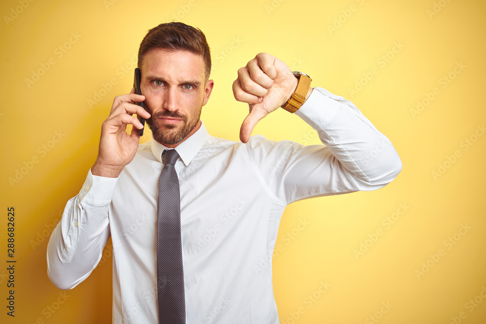 Young handsome business man talking on the phone over yellow isolated background with angry face, negative sign showing dislike with thumbs down, rejection concept Young handsome business man talking on the phone over yellow isolated background with angry face, negative sign showing dislike with thumbs down, rejection concept
