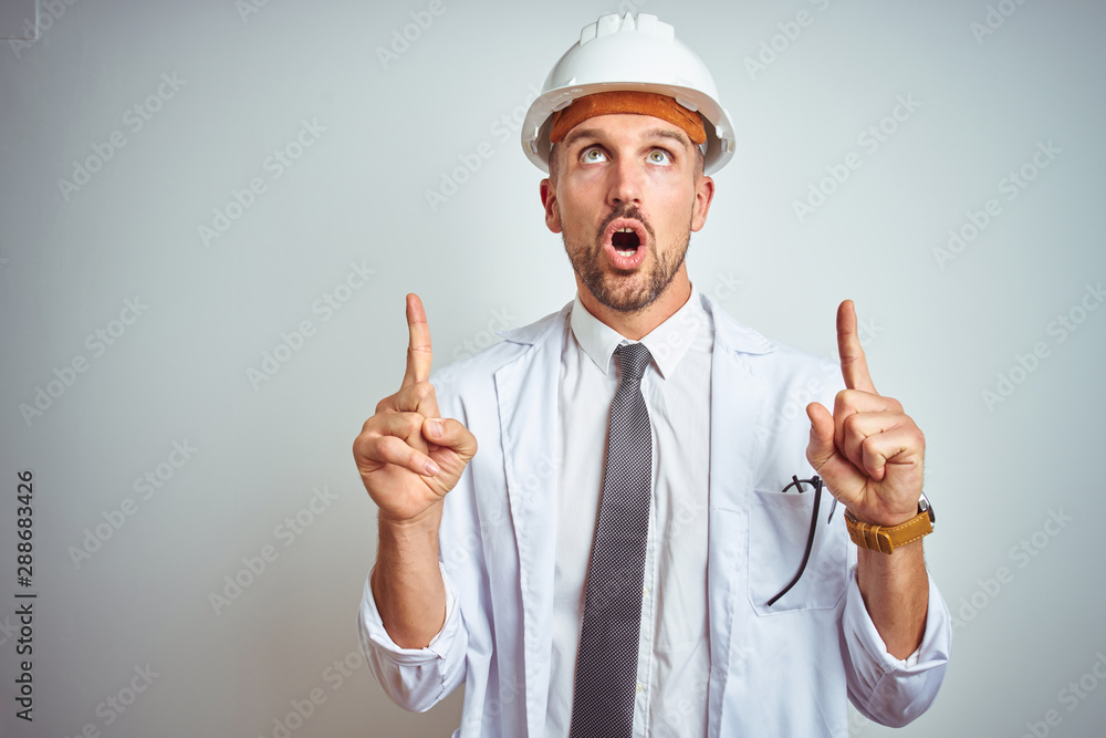 Young handsome engineer man wearing safety helmet over isolated background amazed and surprised looking up and pointing with fingers and raised arms. Young handsome engineer man wearing safety helmet over isolated background amazed and surprised looking up and pointing with fingers and raised arms.