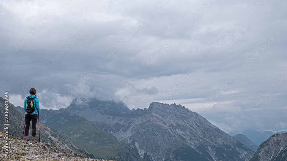 A lone female hiker looks out over some high alpine mountains and valley. Storm clouds gather over the high peaks as she ponders the valley views.