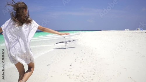 Happy lady in white dress runs after a gray heron along a white sandy beach in the Maldives. A heron flies over the shore. Slow motion. Bora Bora, French Polynesia. Travel motivation.