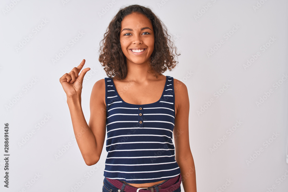 Young brazilian woman wearing striped t-shirt standing over isolated white background smiling and confident gesturing with hand doing small size sign with fingers looking and the camera. Young brazilian woman wearing striped t-shirt standing over isolated white background smiling and confident gesturing with hand doing small size sign with fingers looking and the camera.